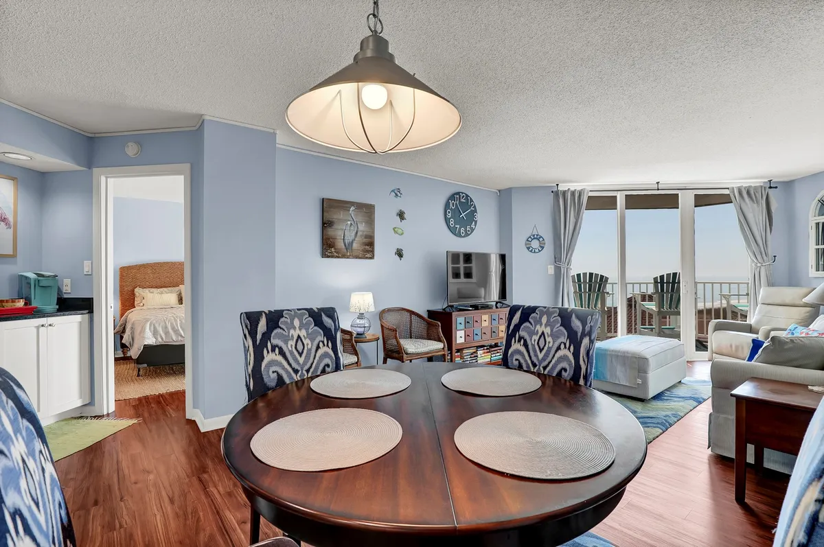 This image features an open-plan living and dining area filled with natural light. It shows a dining table, a cozy sofa, large windows capturing scenic views, and decorative wall art. The kitchen area is visible, showcasing the seamless flow between spaces.