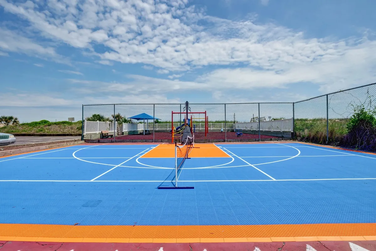 An exterior scene featuring a blue tennis court with orange accents, a net, and a playground visible in the background. The space is designed for tennis, basketball, and children's play, suggesting active outdoor fun for all ages.