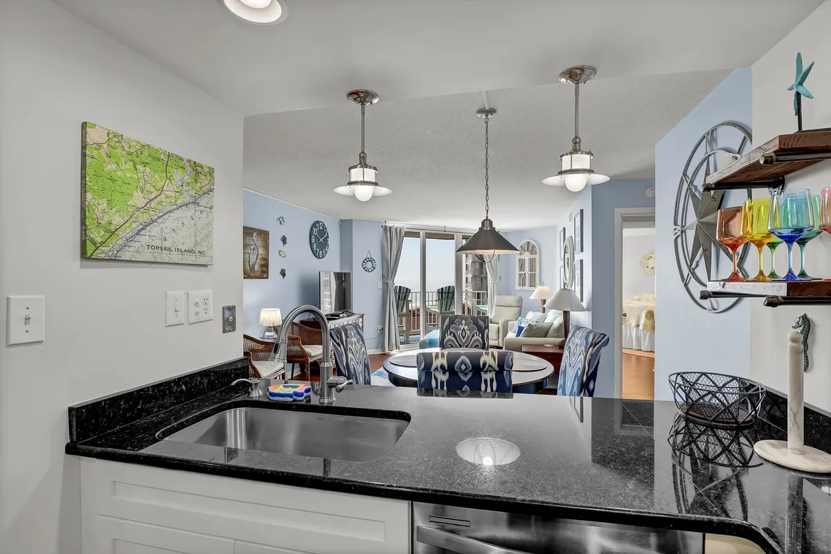 This image focuses on a bright kitchen and dining area with blue chairs, modern pendant lights, colorful glassware, and large windows that frame the ocean view. The kitchen sink and part of the kitchen workspace are clearly visible, emphasizing the space’s functionality for entertaining and preparing meals.