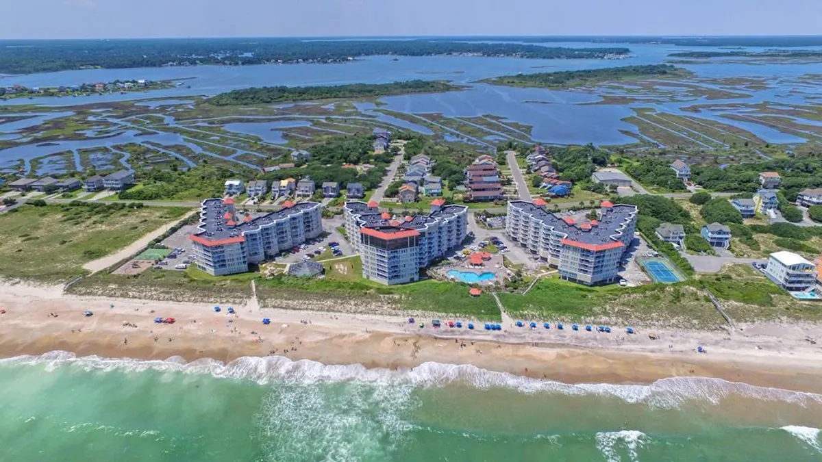 An inviting exterior view showing beachfront buildings, a sparkling swimming pool with pool chairs, umbrellas, and the ocean's waves in the background. The scene suggests easy access from pool to beach and a lively yet relaxed atmosphere.