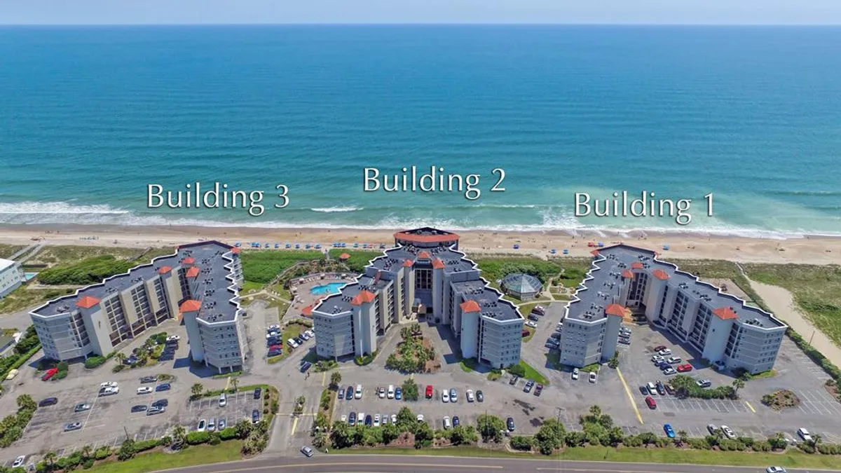 A vibrant outdoor scene featuring three distinctive resort buildings, a sparkling swimming pool, and a glimpse of the ocean beyond. The pool area is lively yet inviting, perfect for both relaxation and social activities.