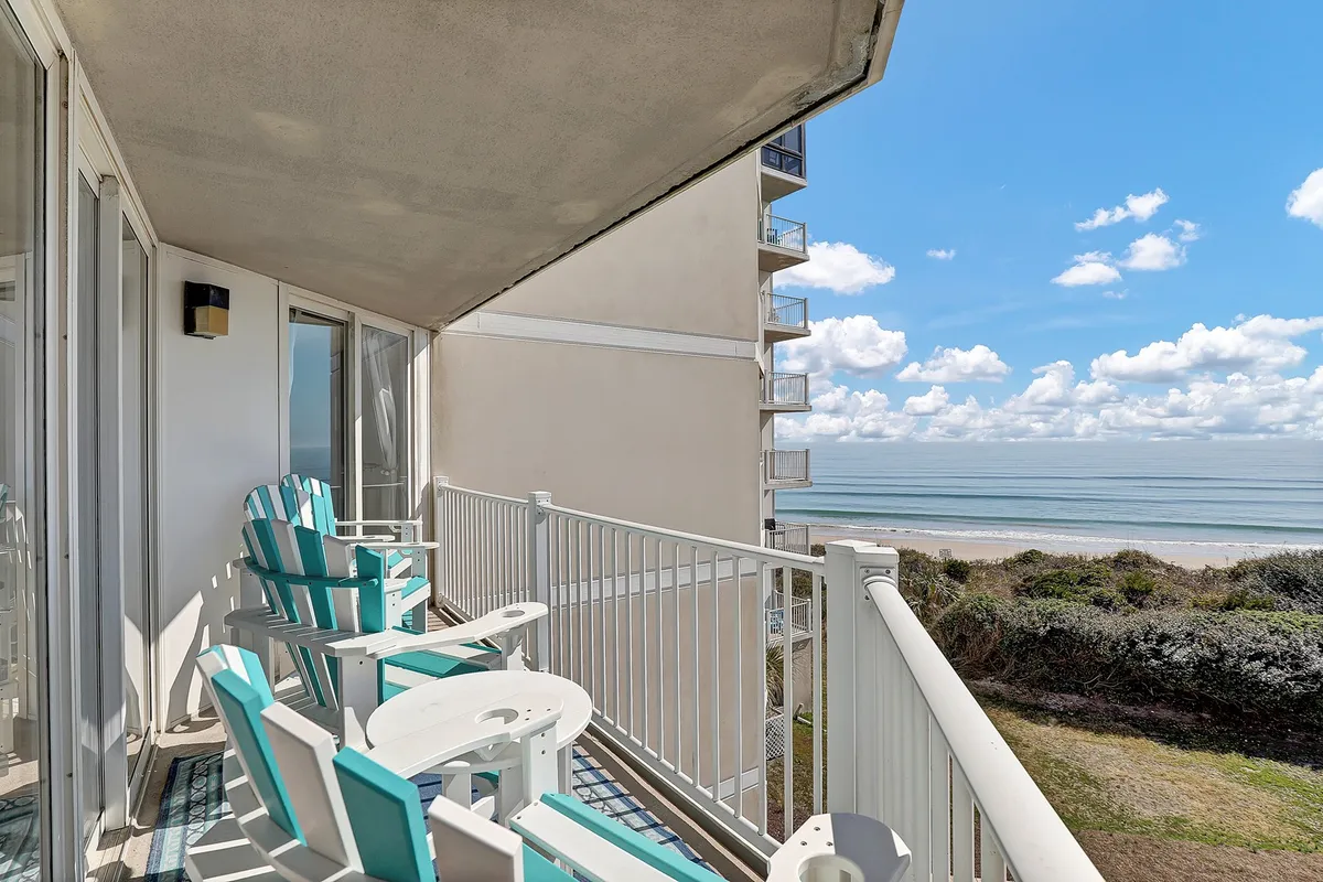 A private balcony setup with comfortable chairs overlooking the ocean, clouds drifting above. The scene is tranquil, perfect for sunrise coffees or sunset relaxation, embodying the indoor/outdoor connection of an oceanfront retreat.