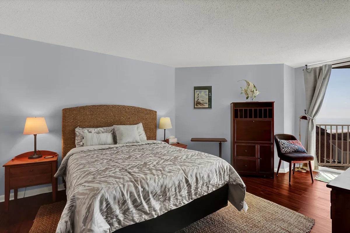 A relaxing bedroom featuring a woven headboard queen bed, flanked by side tables with lamps, a decorative chair, and a large window letting in natural light and offering a scenic view. The room's styling and soft tones exude comfort and tranquility.
