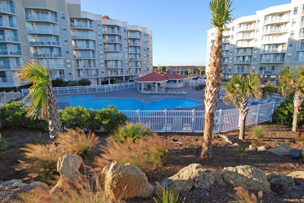A sun-drenched outdoor swimming pool surrounded by landscaped gardens and palm trees, with inviting lounge areas and the architectural facade of the building in the background. The tranquil atmosphere hints at leisurely afternoons spent swimming or relaxing by the water.