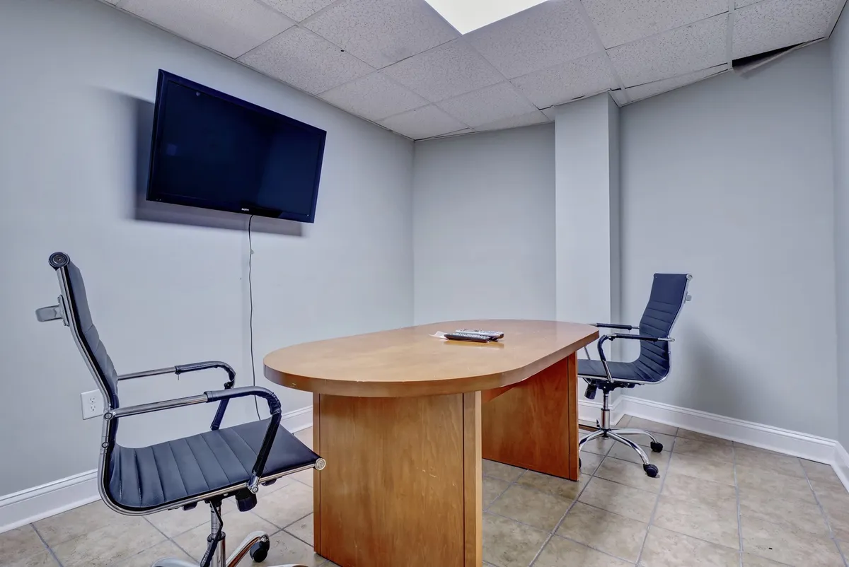A focused work environment featuring a wooden conference table, two office chairs, and a television on the wall. The setup is ideal for business meetings, remote work, or video conferences, emphasizing productivity and convenience.