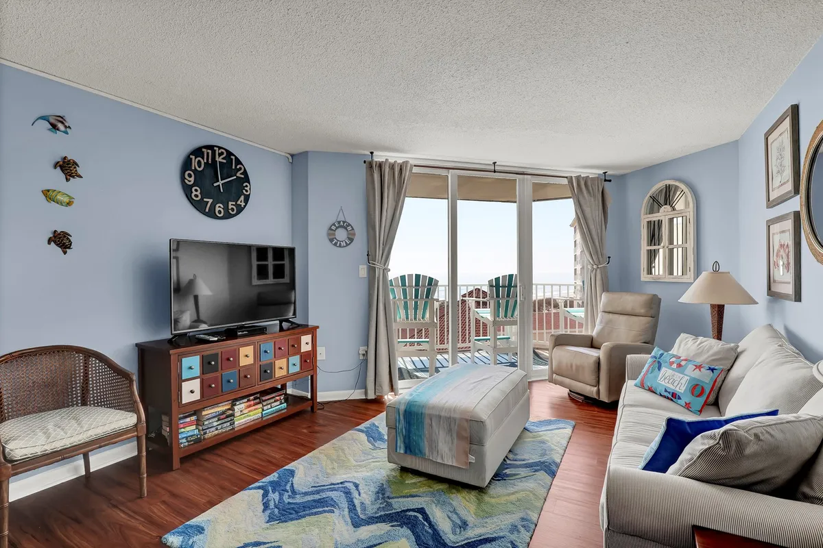 A comfortable living room set-up is shown, with a sofa, coffee table, television, and prominent windows offering an ocean view. The arrangement suggests a space for both relaxation and socializing, with the view connecting indoors to the coastal setting.
